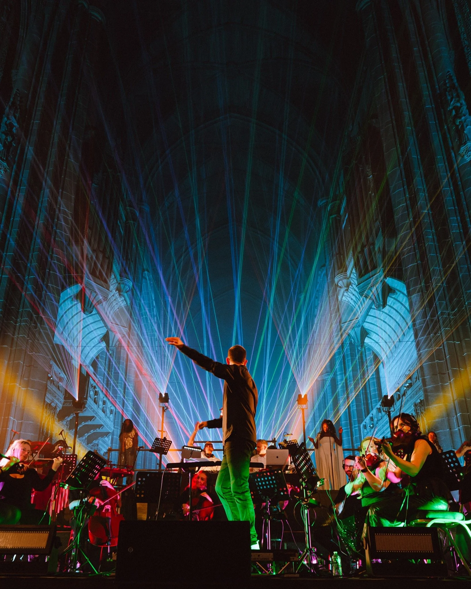 A concert with musicians playing string instruments on stage, a person in the center conducting, colorful laser lights, and a grand architectural interior with high vaulted ceilings.