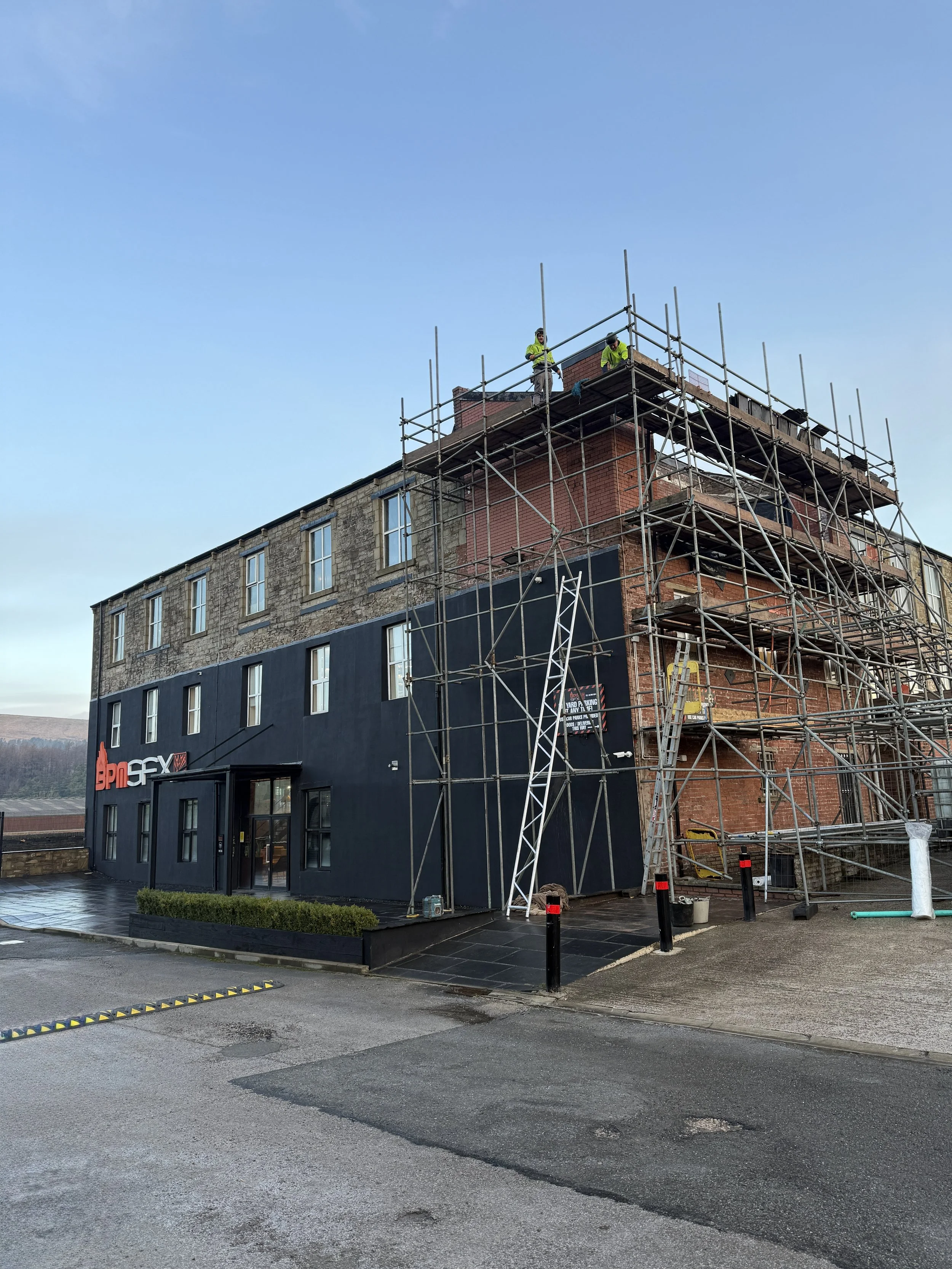 Building under renovation with scaffolding and workers on top, neighboring commercial buildings, wet pavement, cloudy sky.