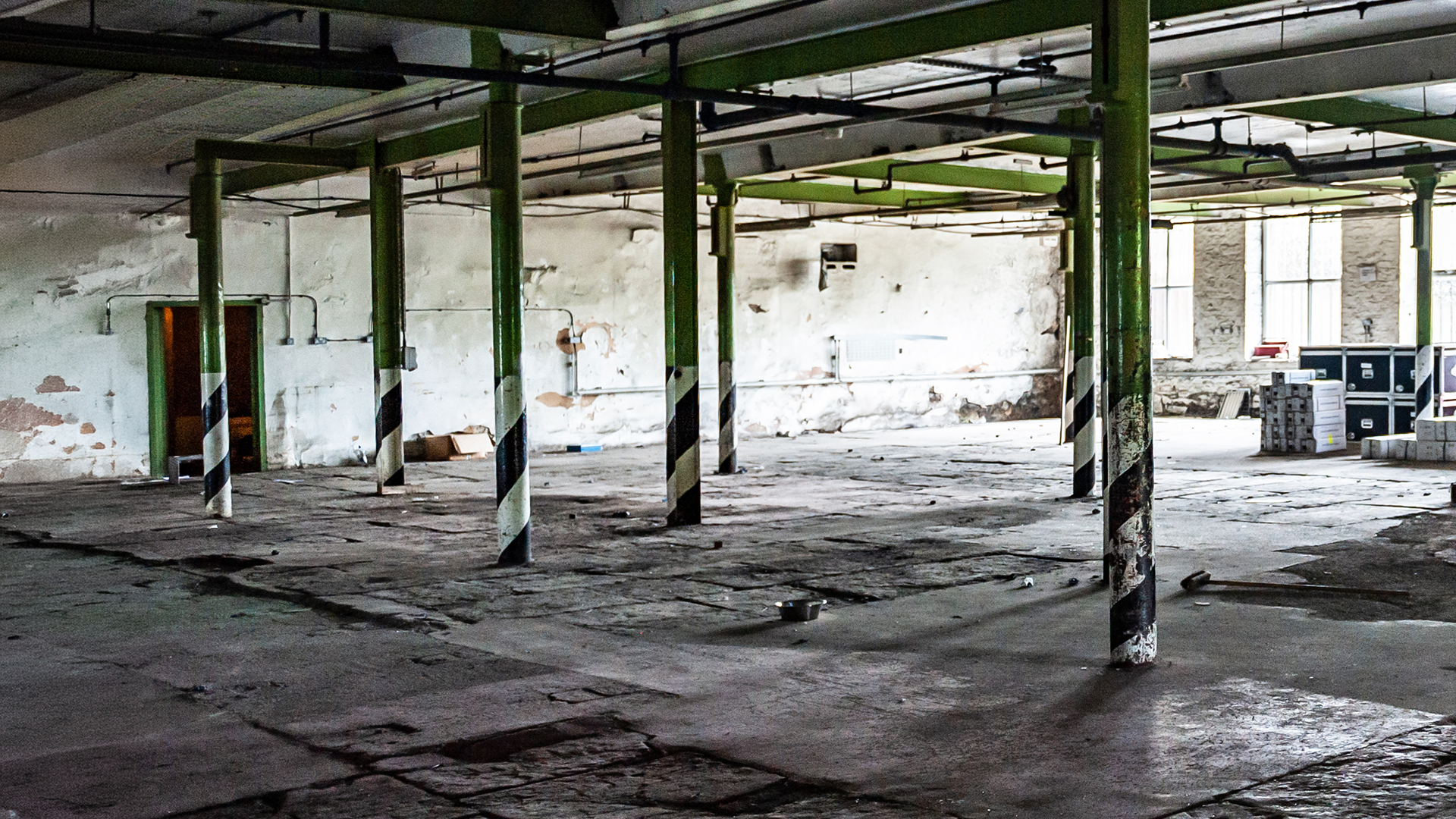 An empty, abandoned, and dilapidated industrial space with peeling paint on the walls, some stacked crates or containers, and exposed pipes and support columns.