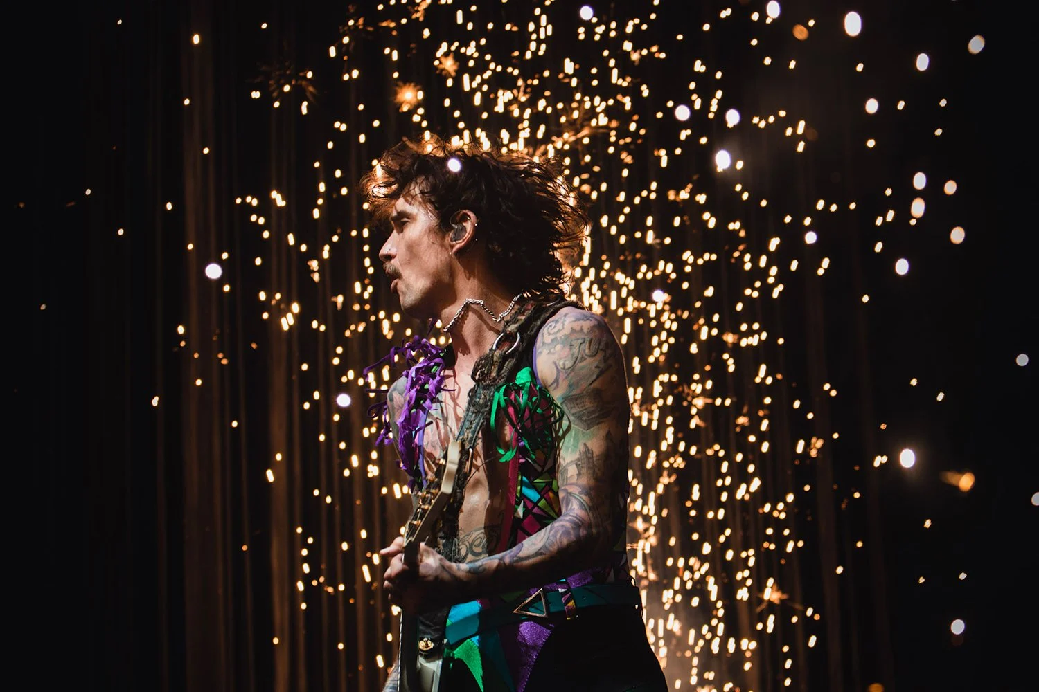 A shirtless male guitarist with tattoos, curly hair, and earrings playing guitar in front of a fiery display of sparklers.