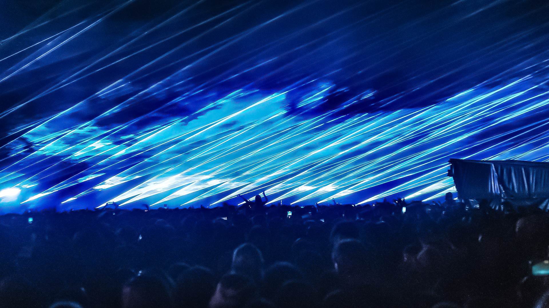 A concert or night event with bright blue laser lights streaming across the dark sky and a large crowd of people in the foreground.