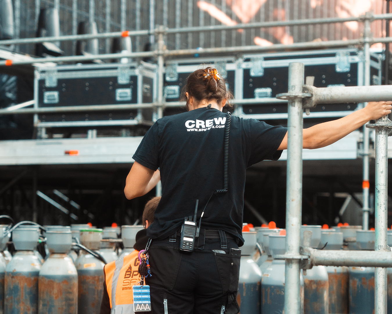 Event staff member wearing a black shirt listed as crew working at a construction or event site, with metal scaffolding and propane tanks in the background.