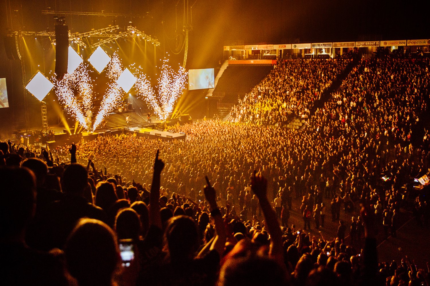 Crowd at a concert with stage lights, pyrotechnics, and large screens
