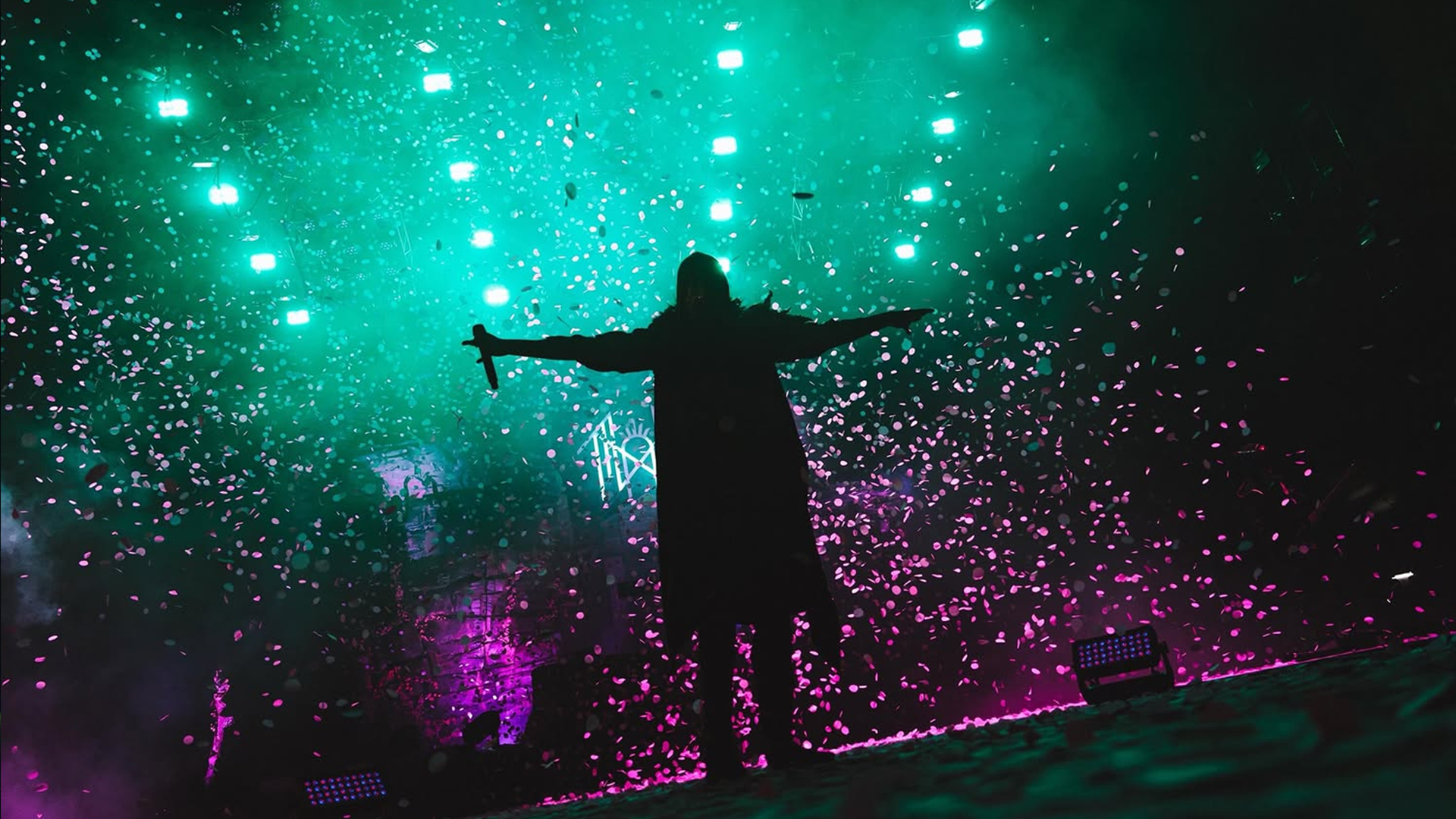 A DJ stands with arms outstretched on a stage illuminated by teal, pink, and purple lights, amidst confetti at a concert or nightclub.