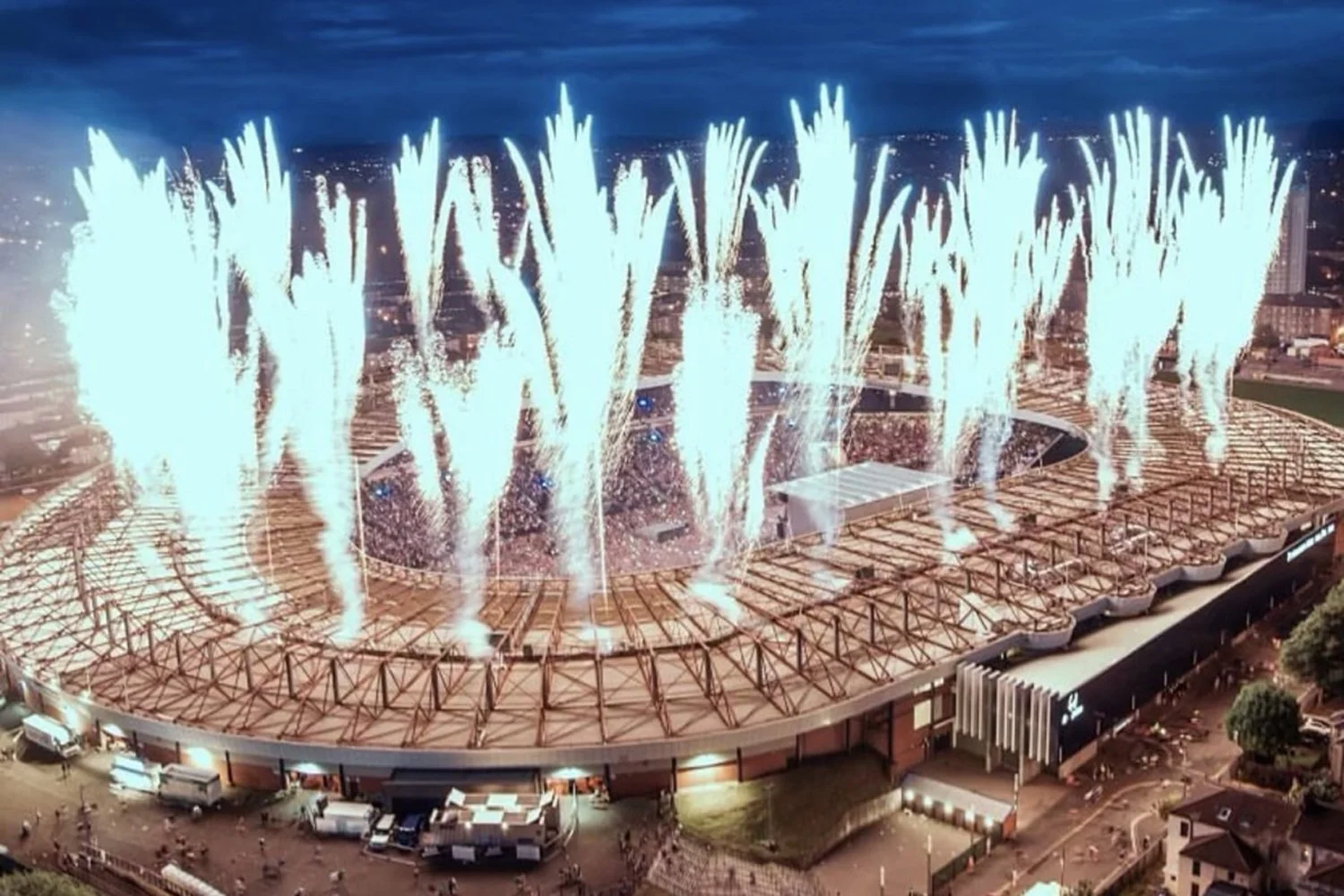 Fireworks display over a sports stadium at night, with bright white explosions lighting up the sky and illuminating the crowd below.