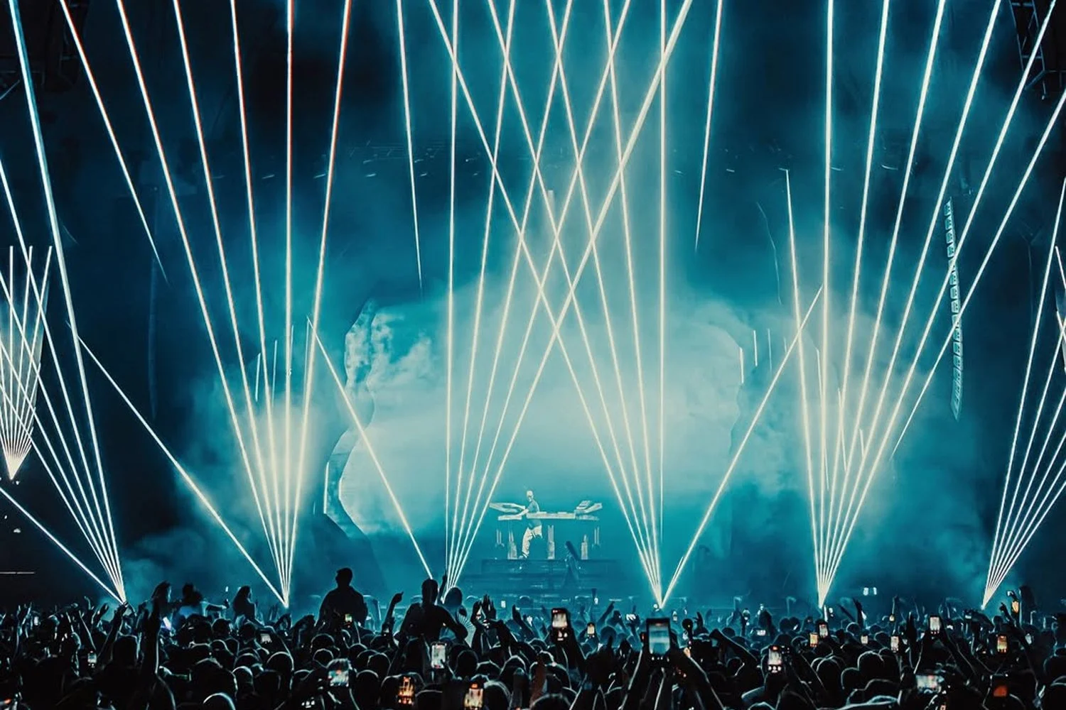 Concert crowd with silhouettes of people taking photos and a DJ performing on stage with laser lights and fog effects.