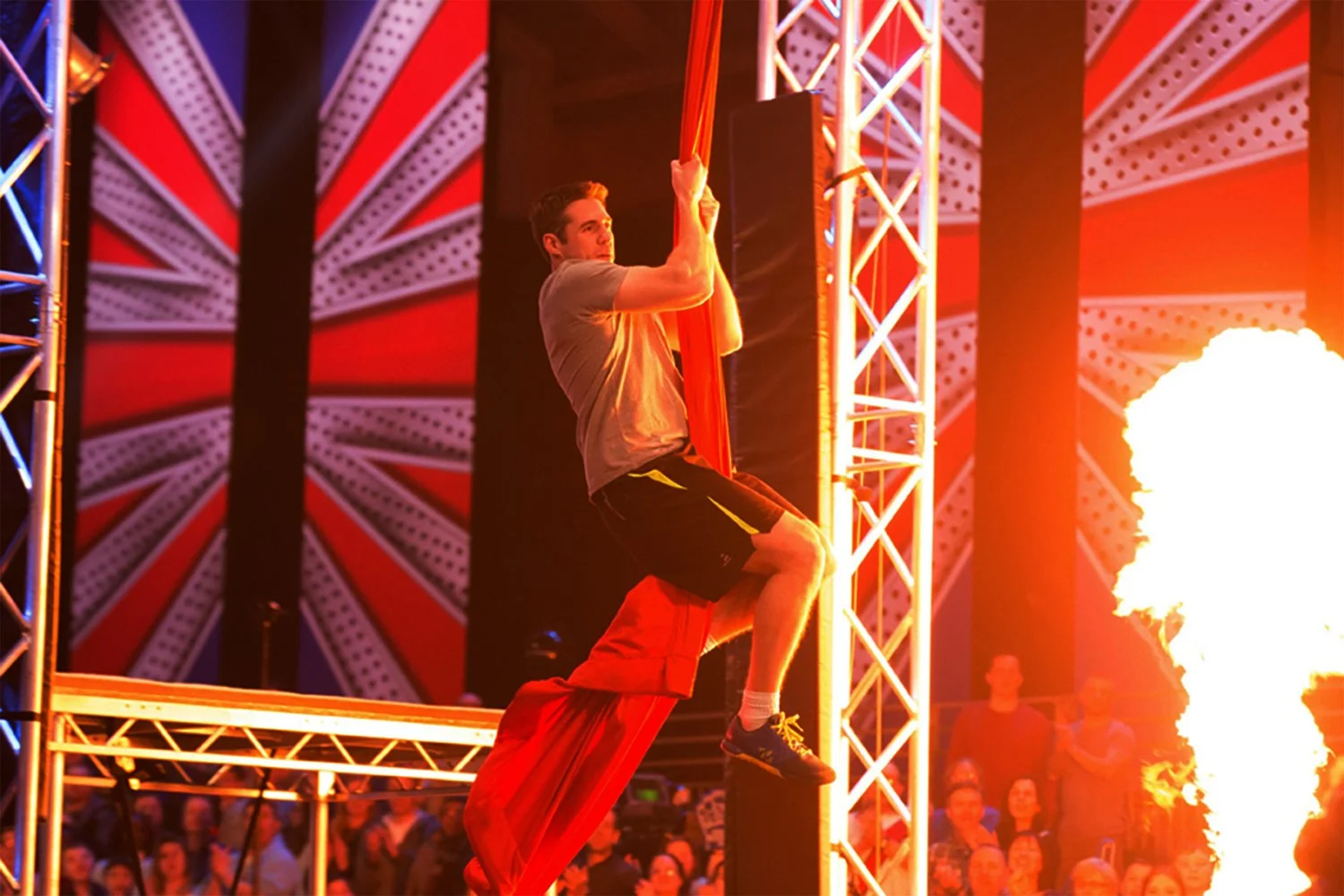 A man climbing an orange aerial silk during a fire performance at a circus or festival, with a large flame on the right and audience members watching in the background.