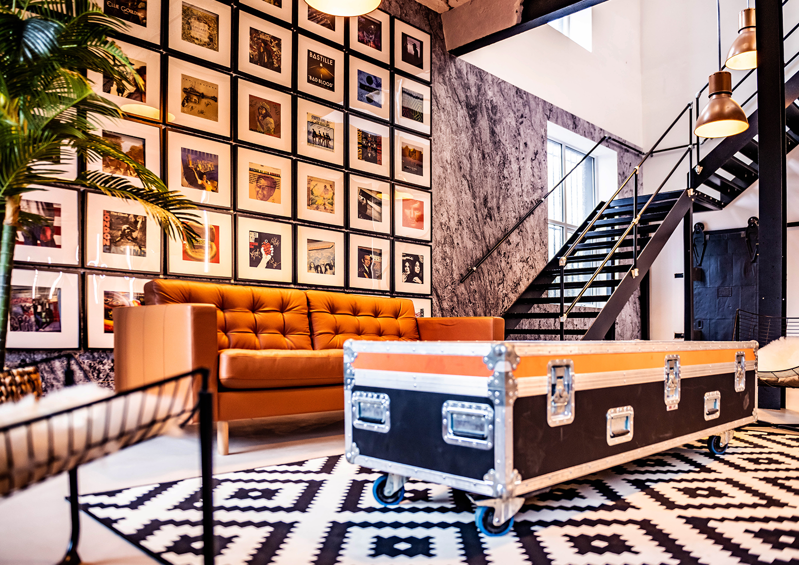 Interior of a stylish living space with a brown tufted sofa, a wall decorated with framed album covers, a tall plant, a black and white patterned rug, and a black metal staircase with pendant lights