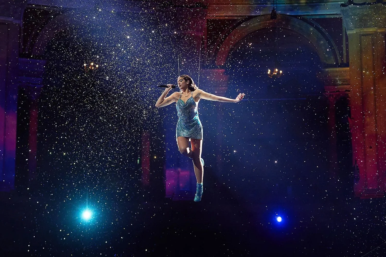 A female singer in a sparkly blue dress performing on stage surrounded by a starry background with lights and arches.