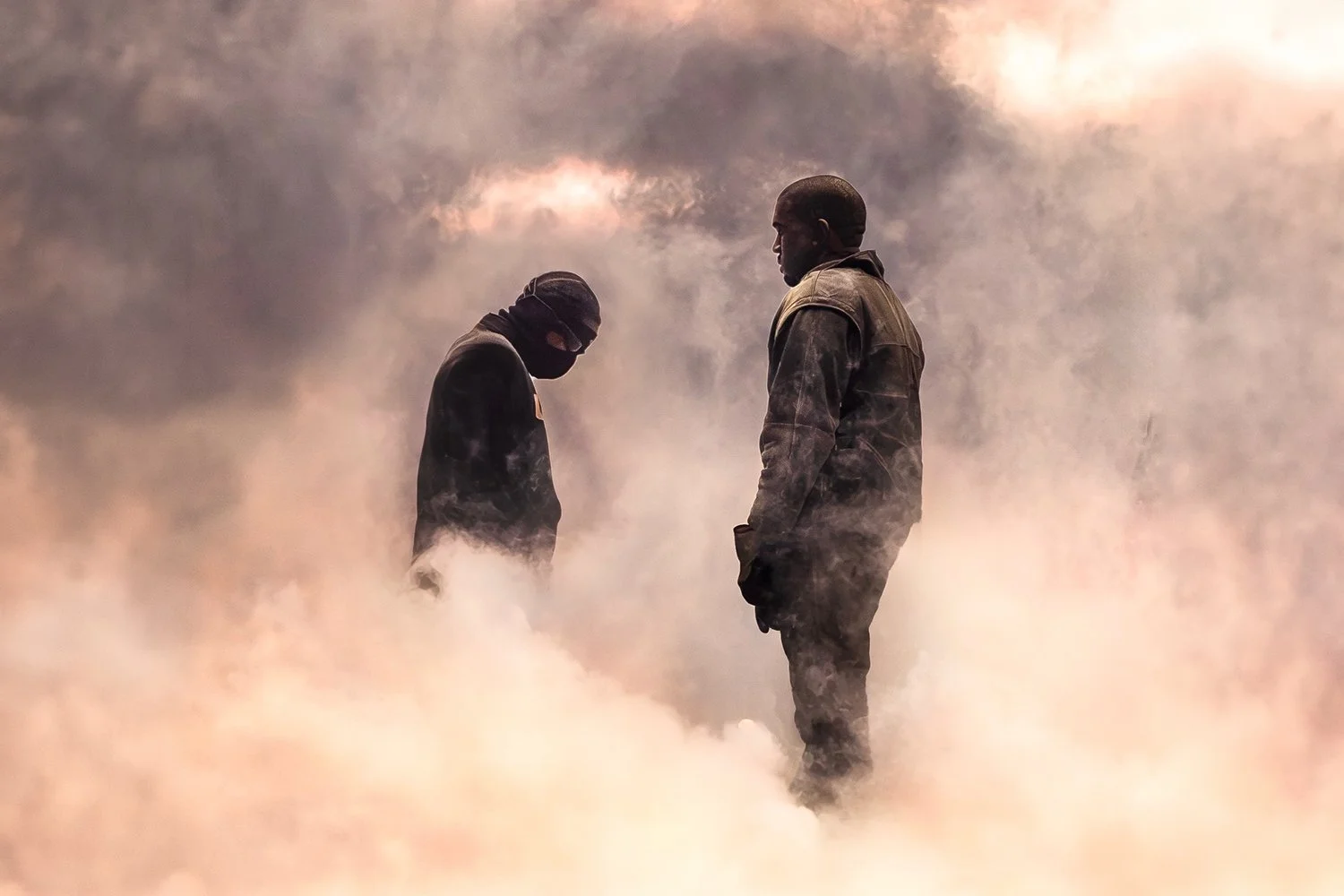 Two men standing amidst a cloud of smoke, one with a mask and the other with a beard, both wearing dark clothing.