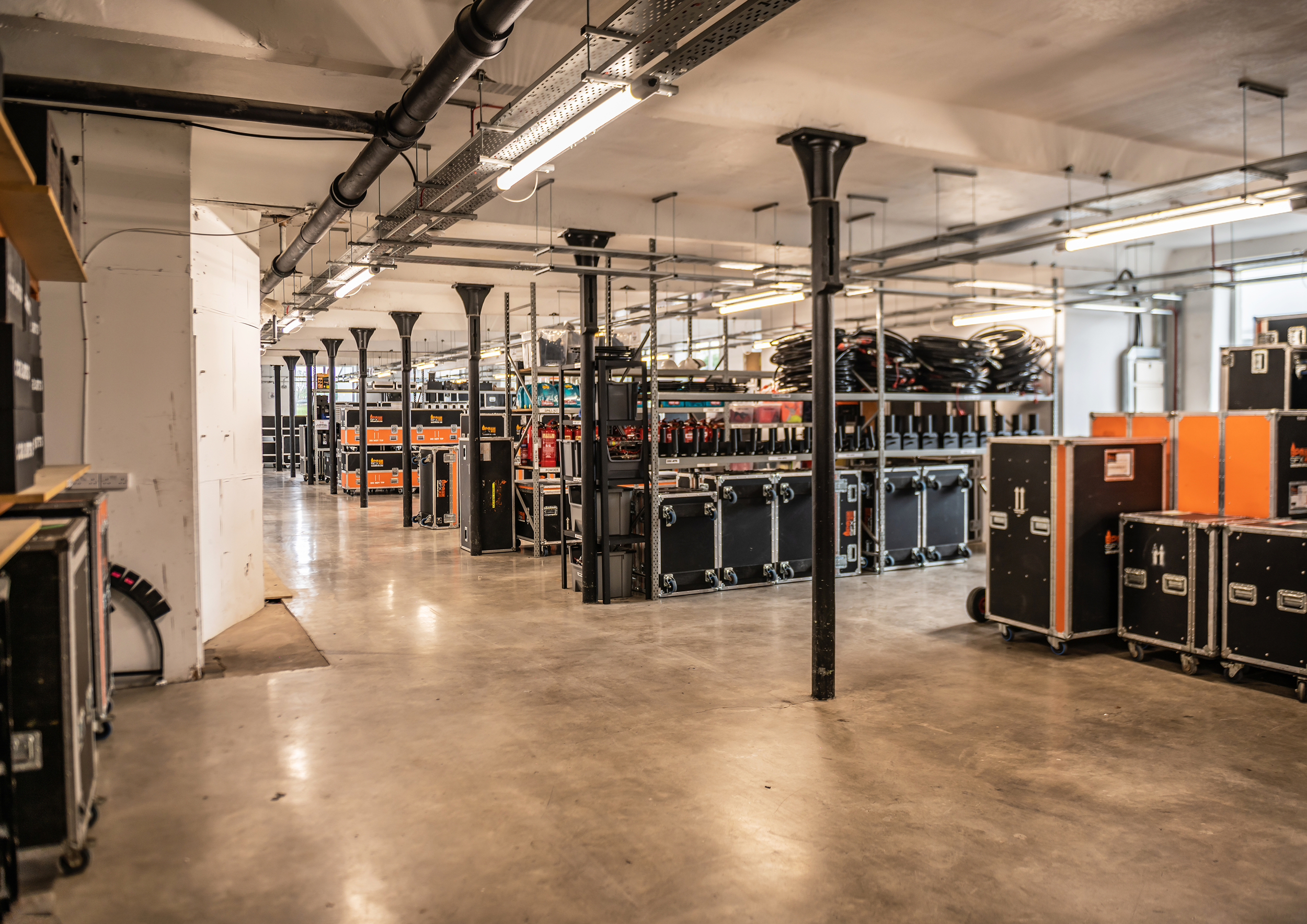 An industrial storage room with black metal shelves holding various equipment, electrical boxes, and coiled cables. The room has a concrete floor, white walls, and exposed ceiling pipes and lighting fixtures.