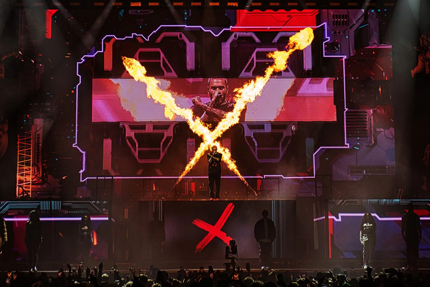 A performer on stage with a large digital screen behind them displaying an image of the same performer. The performer is shooting flames outward in an 'X' shape, with an audience in front of the stage.