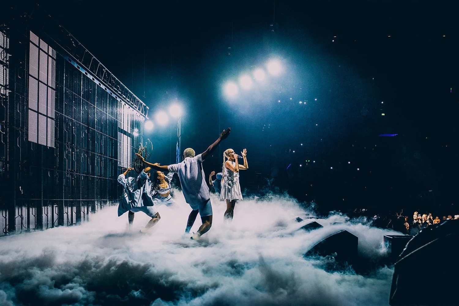People performing on stage with clouds of smoke and bright stage lights, audience visible in the dark background.