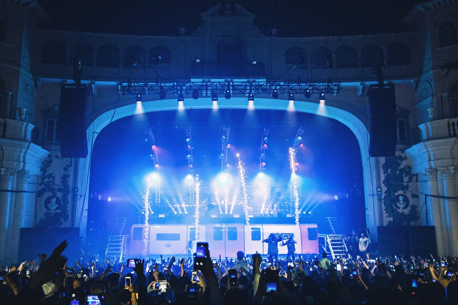 Concert stage with bright blue lights, pyrotechnics, and a large crowd of people taking photos and videos with their phones at night.
