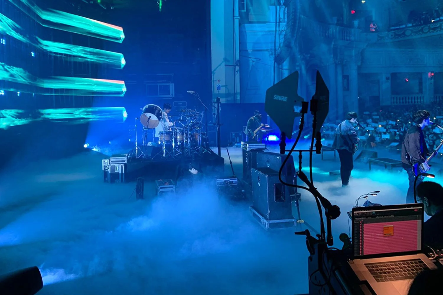 Concert stage with musicians, fog effects, and blue lighting, seen from the side with a sound technician working on a laptop in the foreground.