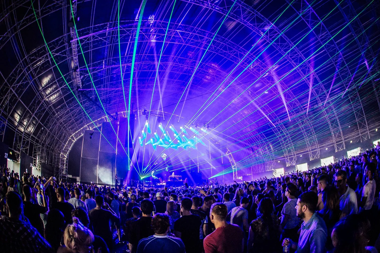 Crowd attending a live electronic music concert inside large, arched stadium with colorful laser light show.