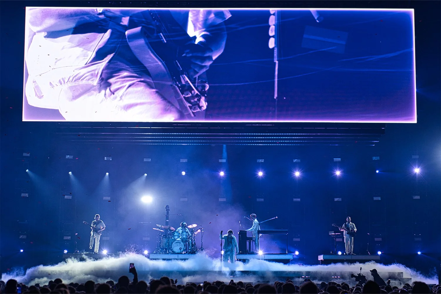 A live music concert taking place with a band performing on a stage illuminated by blue and purple lights, a large screen displaying a close-up of the guitarist, fog effects on the floor, and an audience in the foreground.
