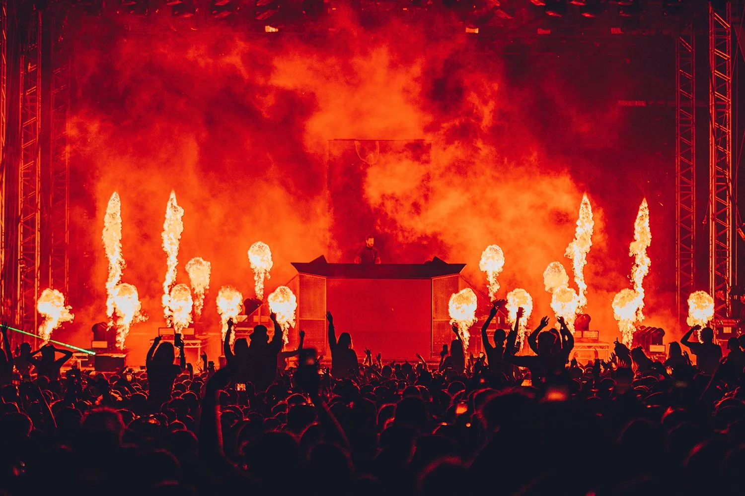 A concert with a DJ on stage surrounded by flames and pyrotechnics, with a large crowd of people dancing and raising their hands.