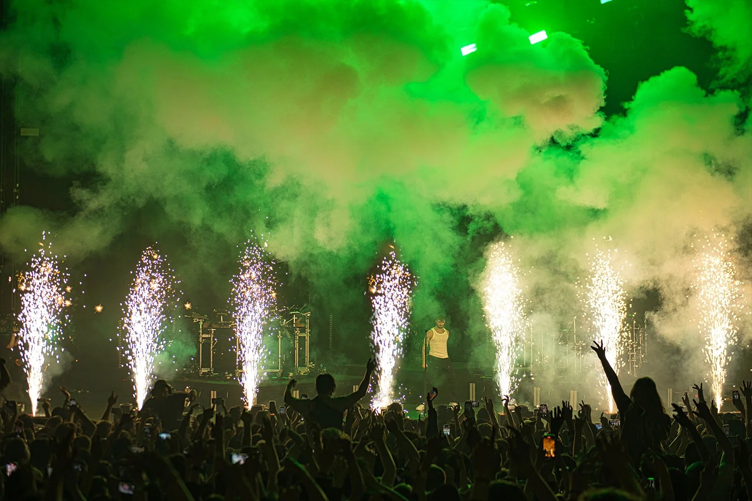 A concert scene with a performer on stage, surrounded by green and white smoke, pyrotechnics, and an audience with raised hands and phones.