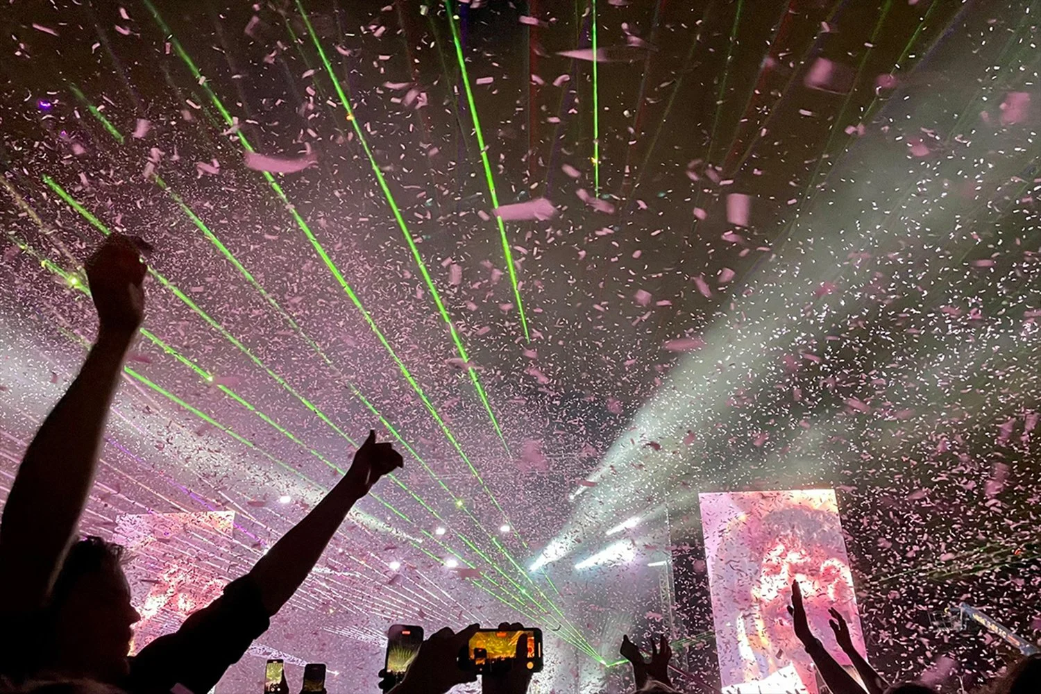 Concert scene with a crowd of people raising their hands, confetti falling, and green laser lights shining from the stage.