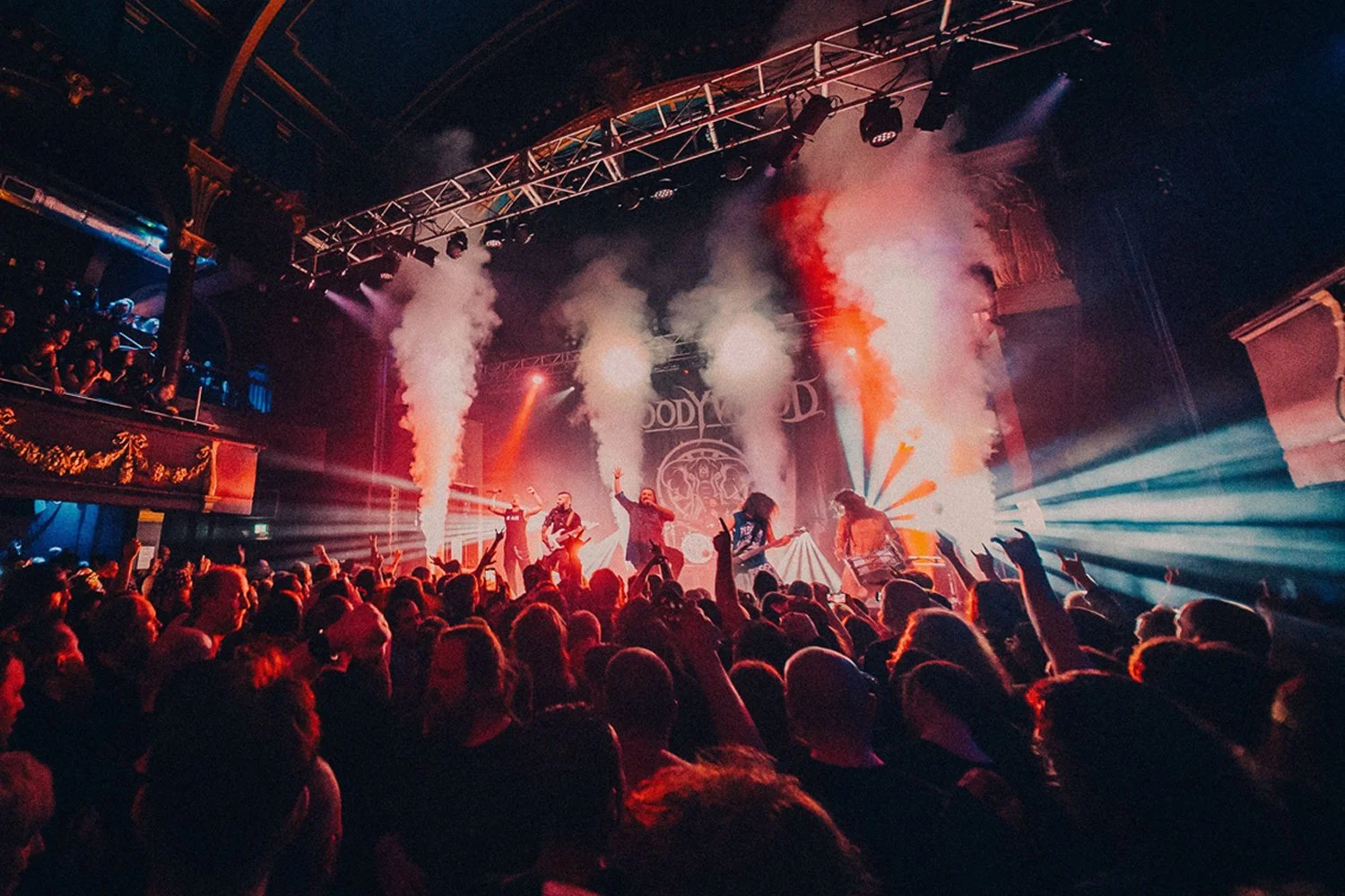 A concert with a band performing on stage, smoke effects, colorful lighting, and a crowd watching and raising their hands.