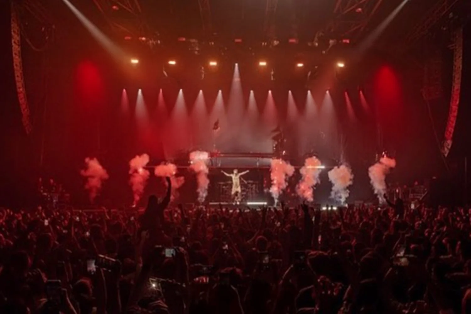 A crowded concert with a singer performing on stage, with red and orange lighting, smoke clouds, and an audience raising their hands.