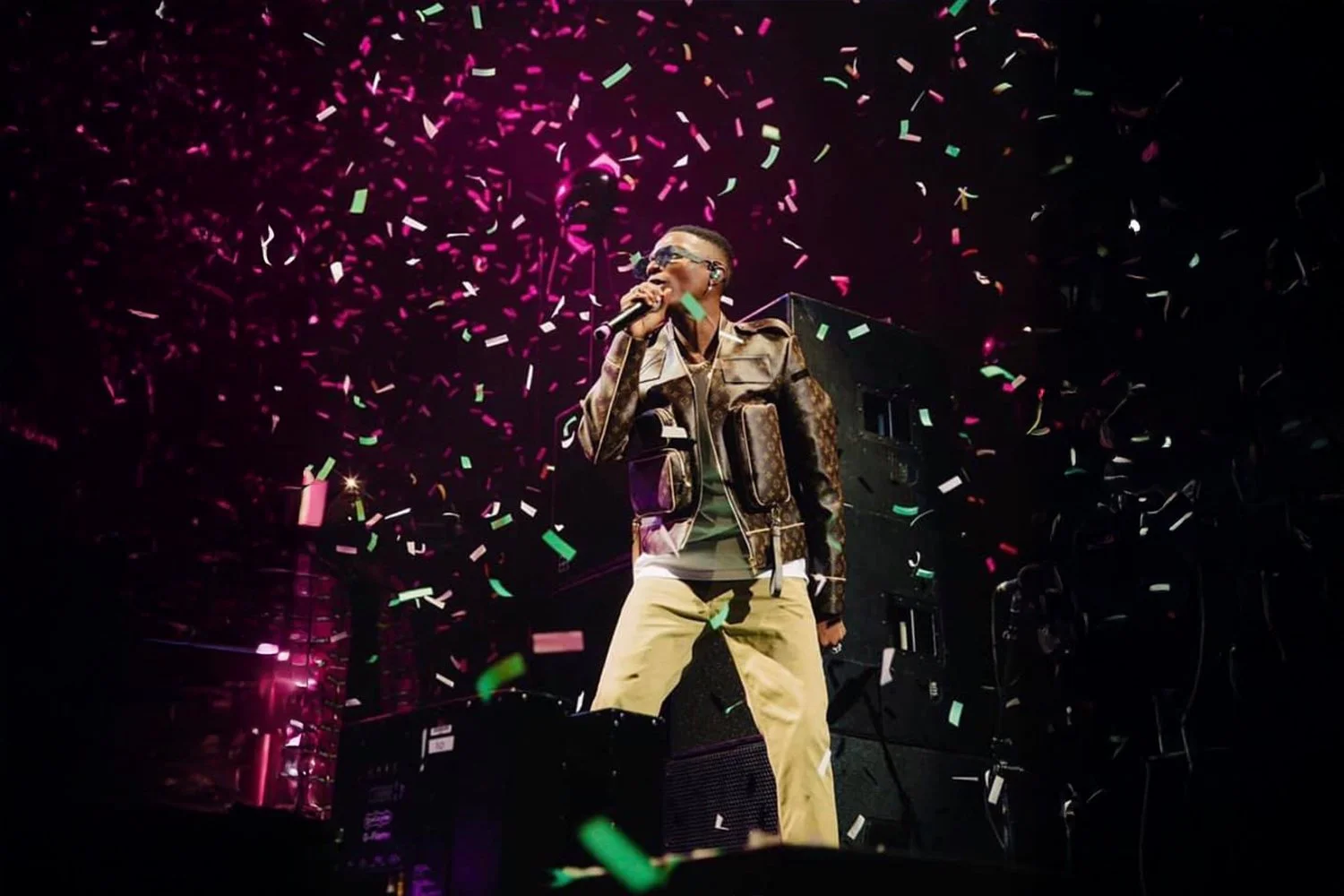 A singer performs on stage in front of colorful confetti, wearing sunglasses, a patterned jacket, and yellow pants, with a microphone in hand.