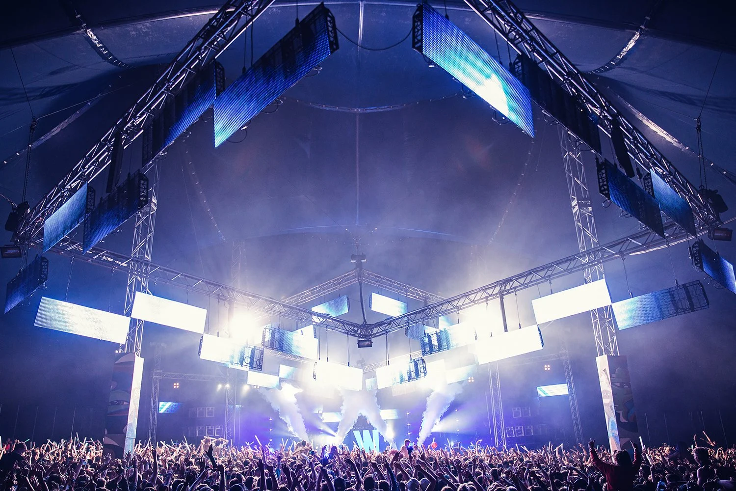 A large concert or music festival with a stage surrounded by multiple LED screens and lighting equipment, and a crowd of people with raised hands enjoying the live performance.