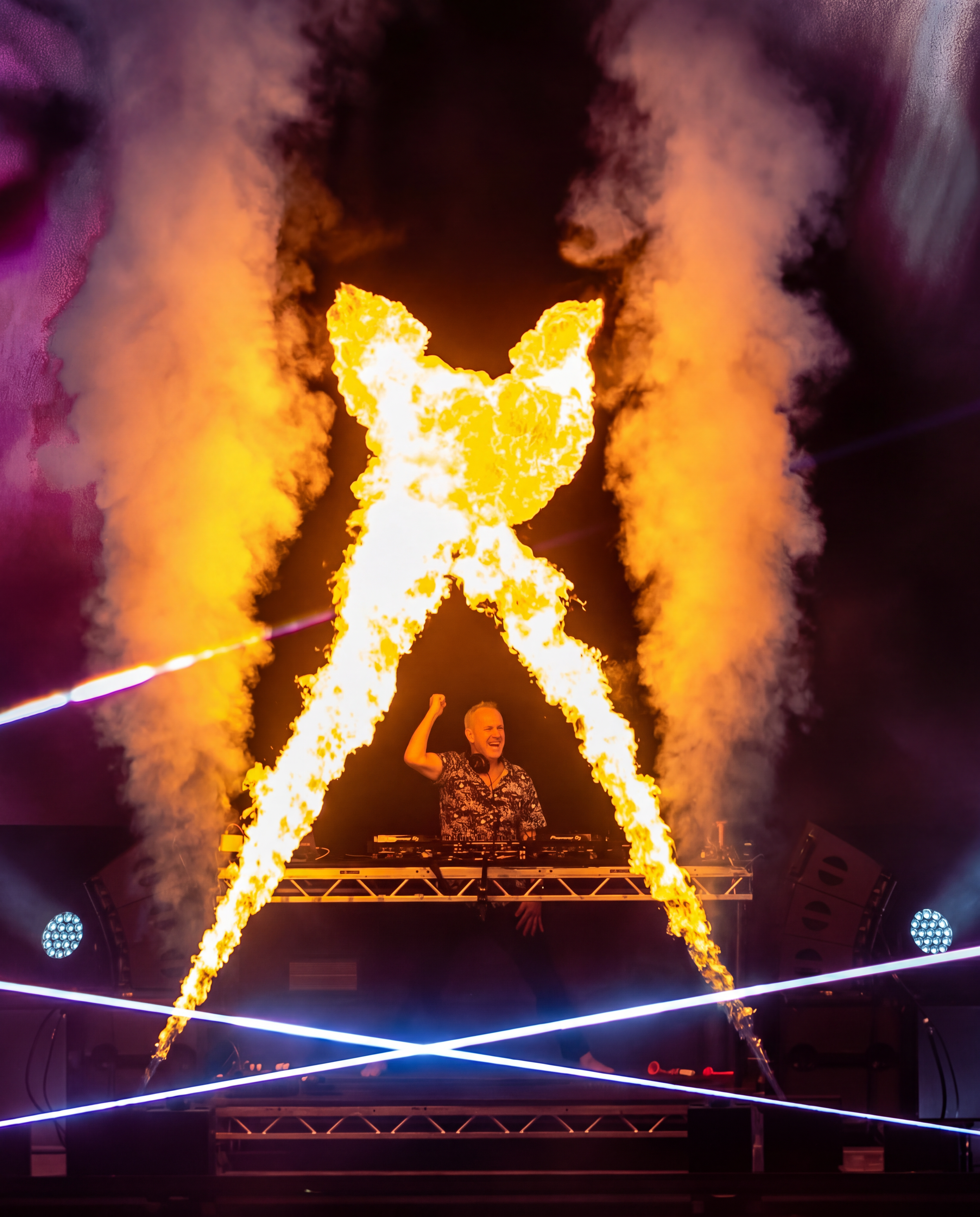 A DJ stands behind a console with fire and smoke effects at a music event, smiling and raising one arm, with colorful stage lighting surrounding him.