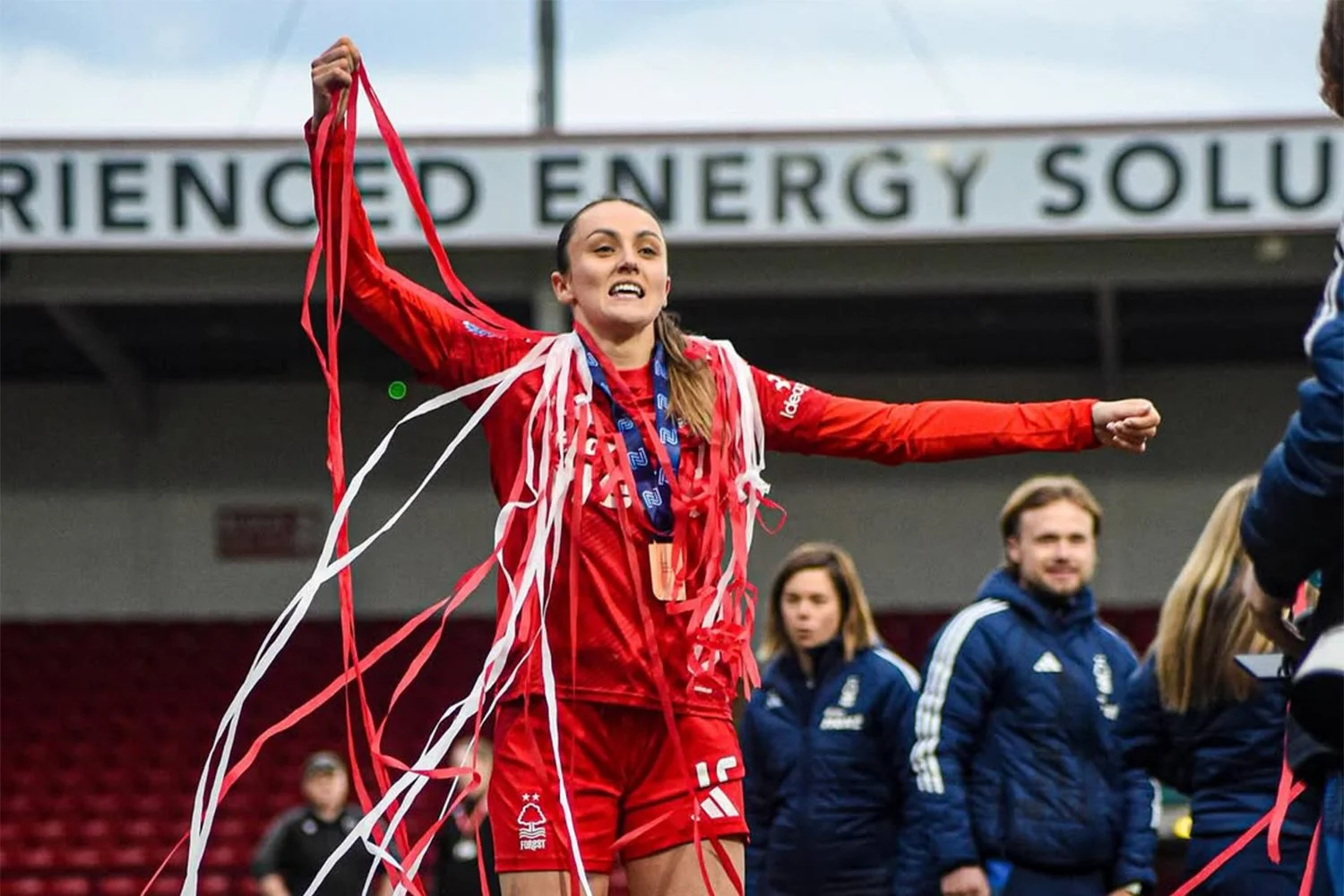 A female athlete in a red sports uniform with a Nottingham Forest logo celebrates victory, with red and white streamers on her. She has medals around her neck and is on a stadium field, with other team members and officials in the background.