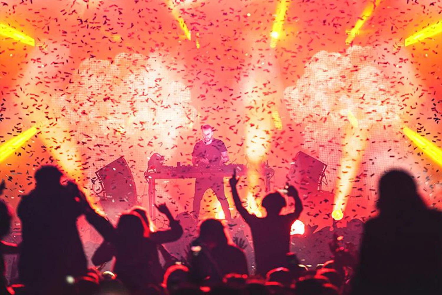 A DJ performing on stage with colored smoke, confetti, and bright lights while an audience dances and enjoys the concert.