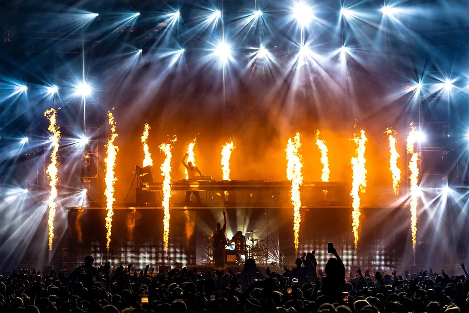 A concert scene with bright stage lights, pyrotechnics shooting fire upwards, and a DJ or performer on stage surrounded by an audience taking photos.