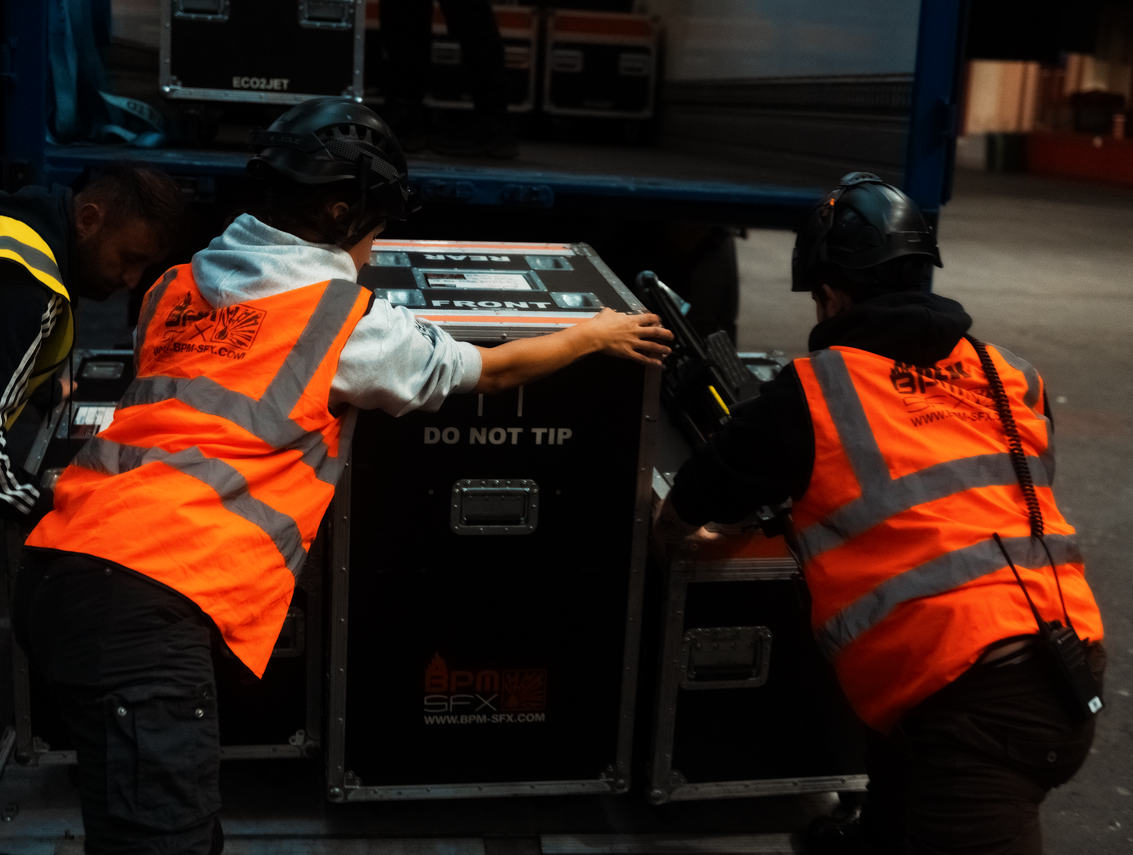 Three workers wearing safety helmets and orange vests are handling a large case labeled 'FRAGILE' and 'DO NOT TIP' on a truck or storage area.
