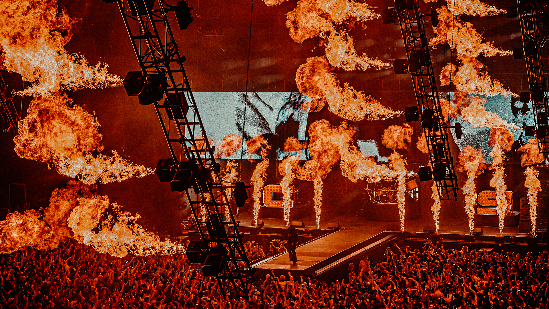A concert stage with performers, surrounded by pyrotechnics and flames, audience in front, and stage lights overhead.