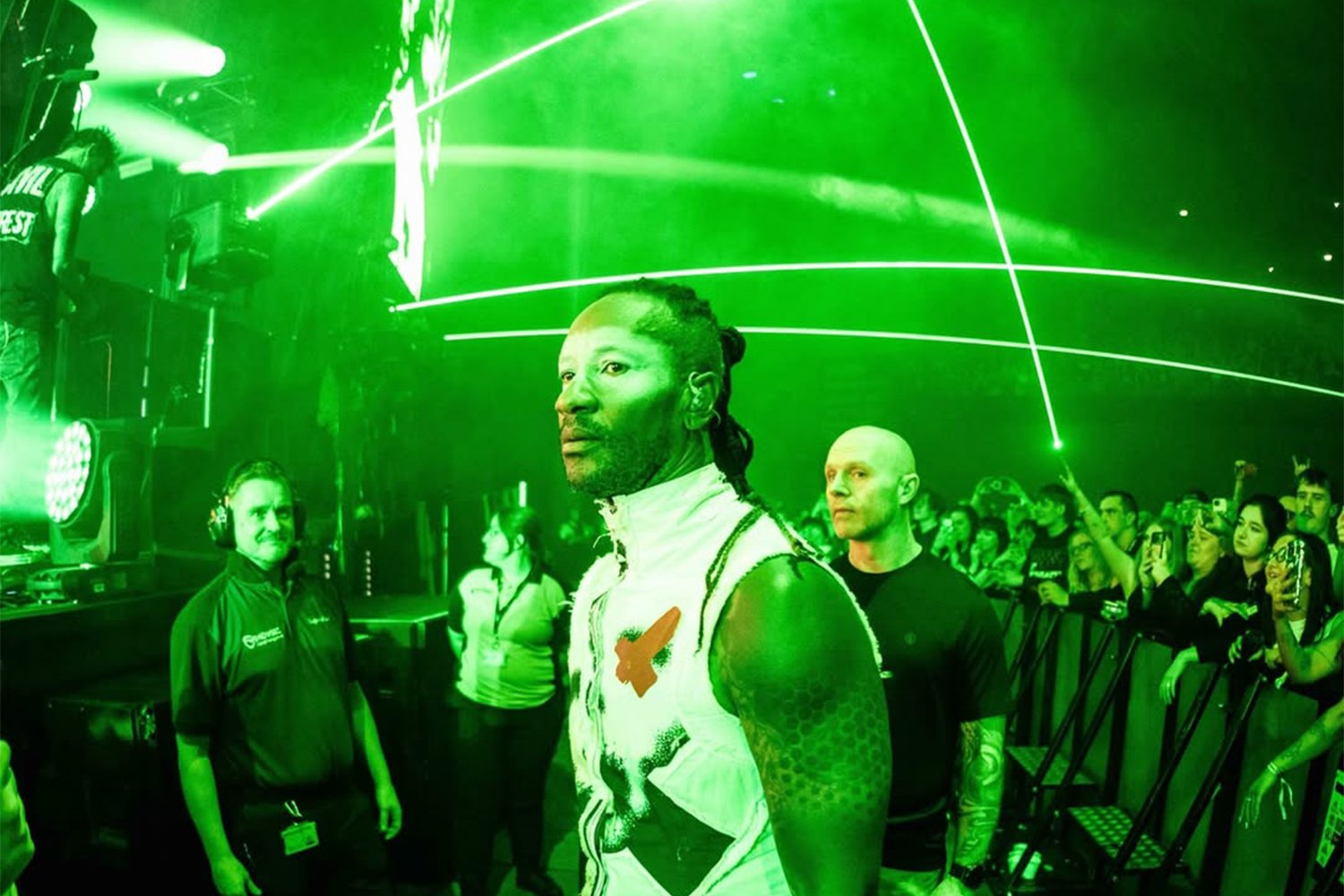 A man with a serious expression, sporting a sleeveless white top with a graphic on the front, stands at a party or music event with green laser lights overhead. A crowd is visible in the background, along with a DJ on the left side near the sound equipment.