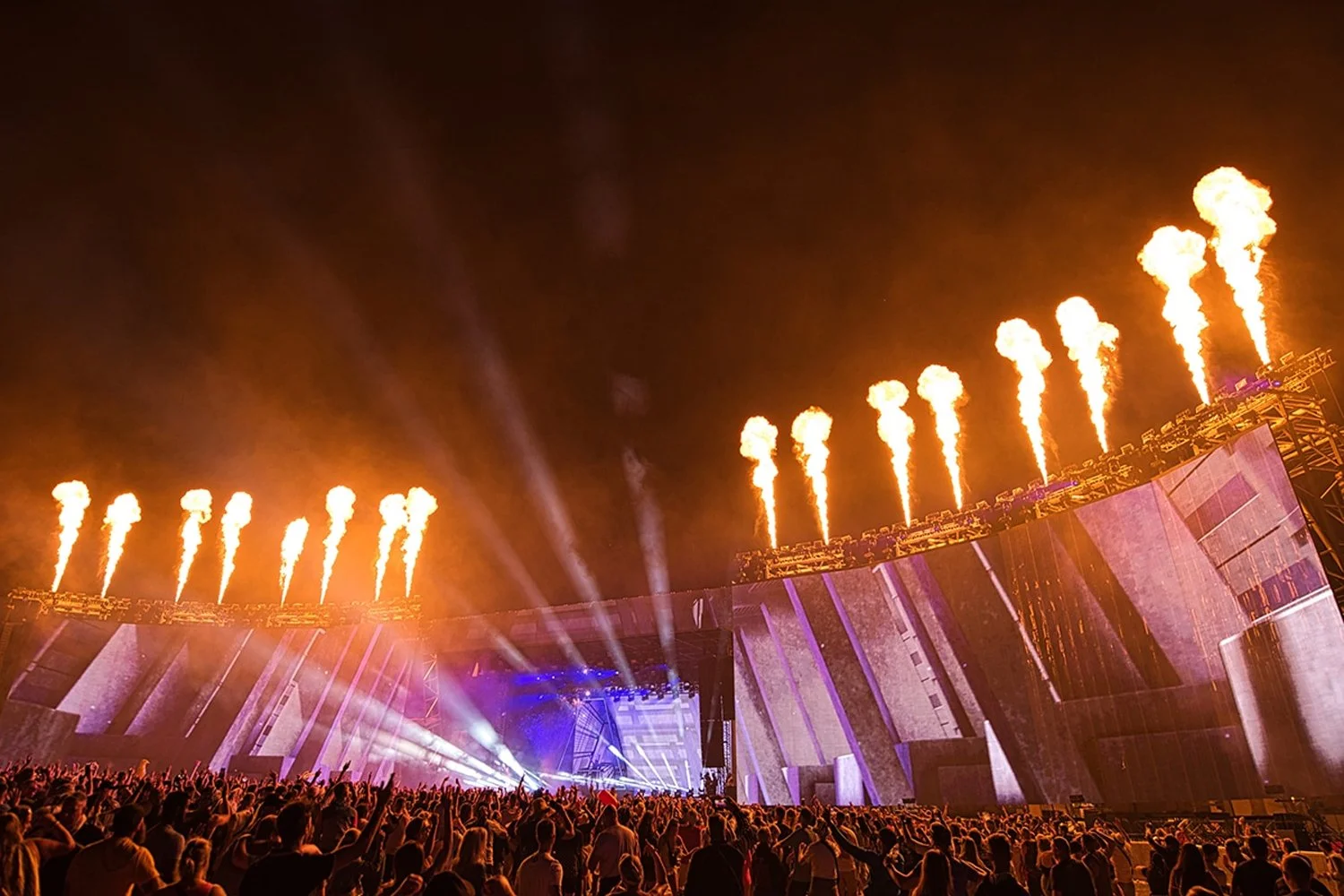 A large crowd watching a nighttime outdoor concert with pyrotechnics and bright stage lights.