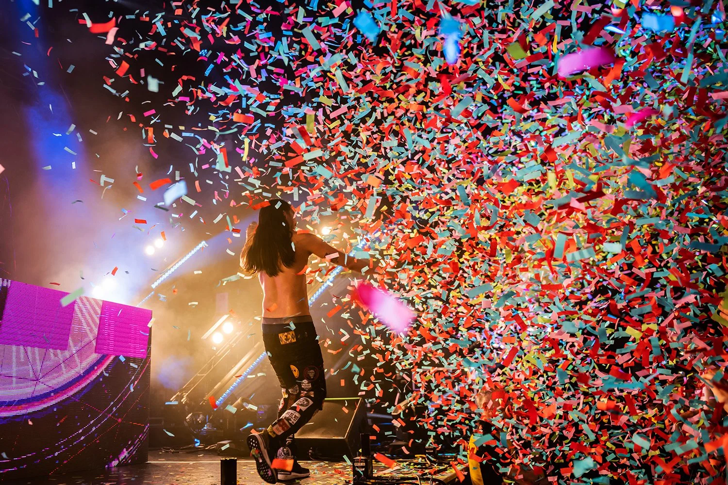 A performer on stage with long hair, shirtless, wearing patched pants, surrounded by colorful confetti and stage lights.