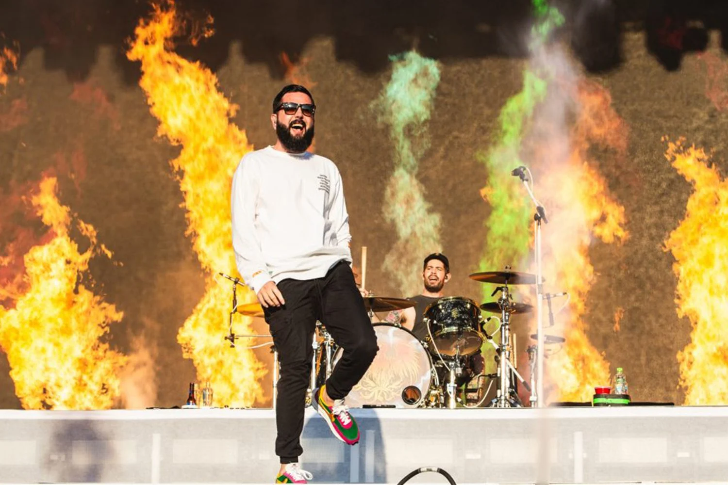 A man singing and dancing on stage with a drummer behind him, flames and colorful smoke in the background.