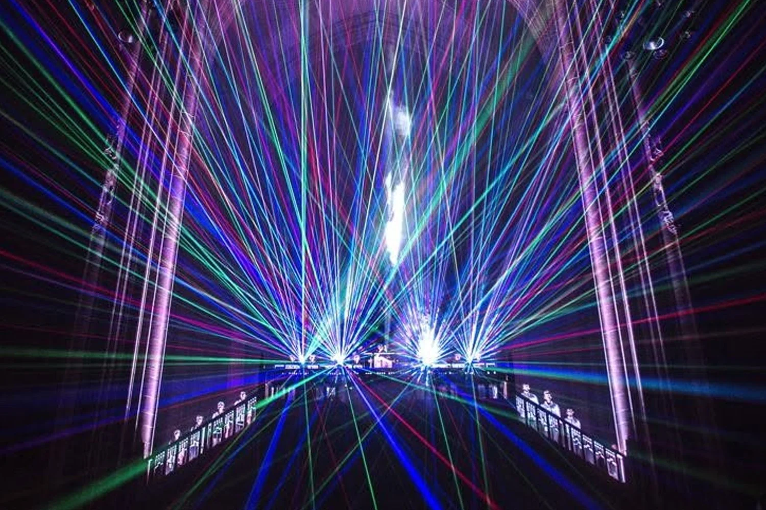 A colorful laser light show at night with beams of green, blue, purple, and red illuminating a structure that resembles a bridge or archway.