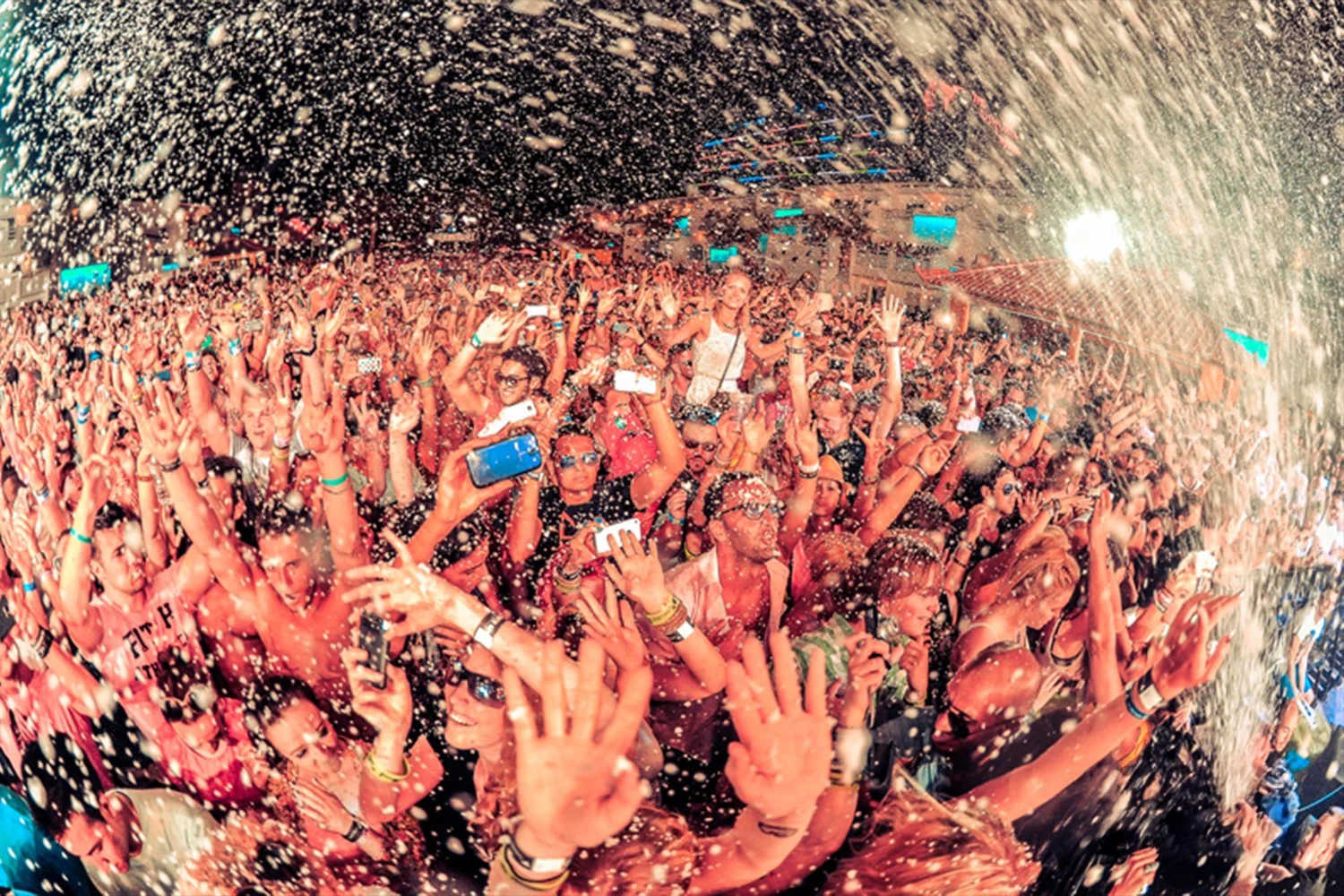 Crowd of people at a concert or festival with foam and water being sprayed over them, some holding up phones to take pictures, with vibrant lighting.