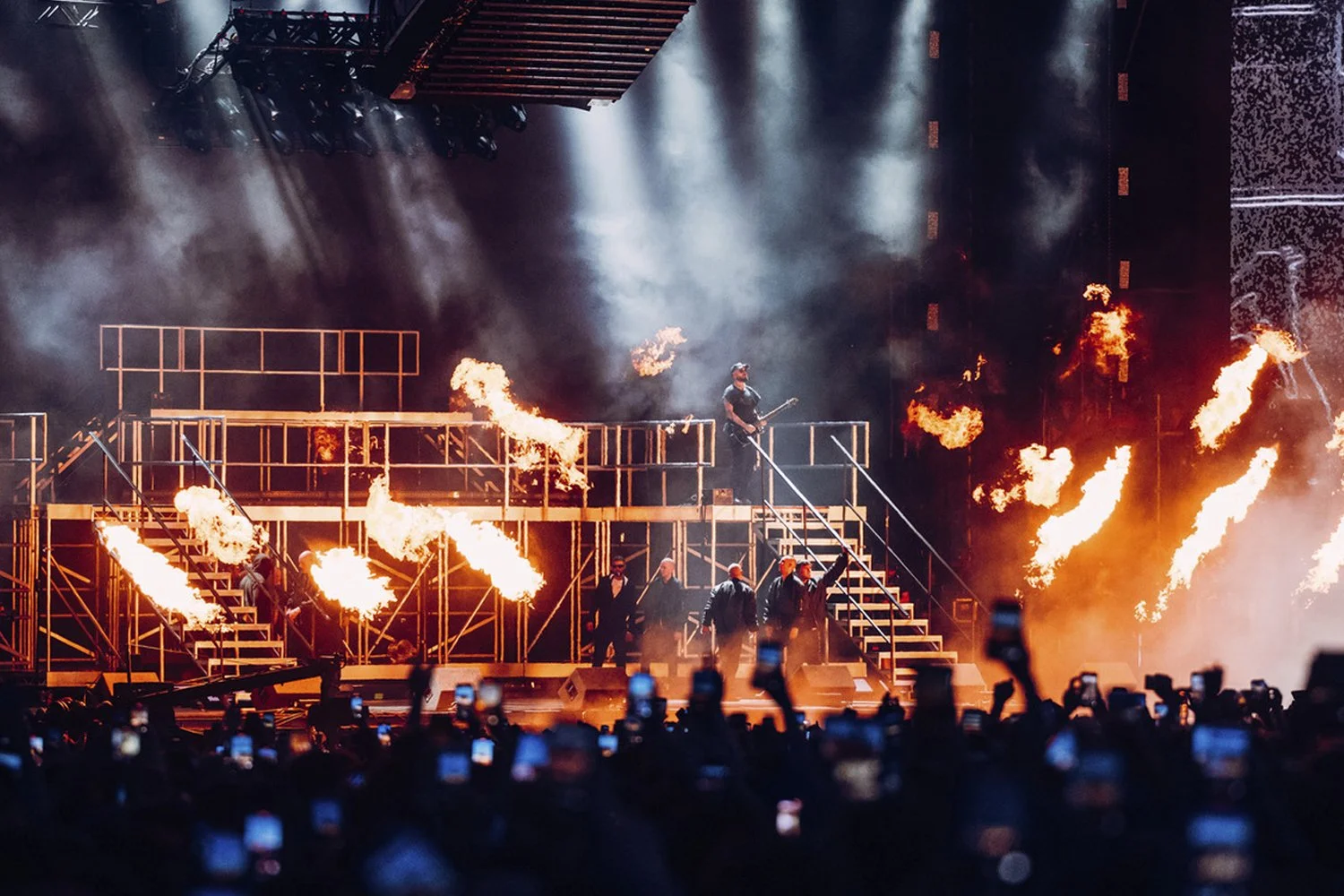 A stage performance with fire effects, featuring a person playing guitar on an elevated platform surrounded by fire explosions, with smoke and stage lighting in the background, and an audience in front capturing the moment.