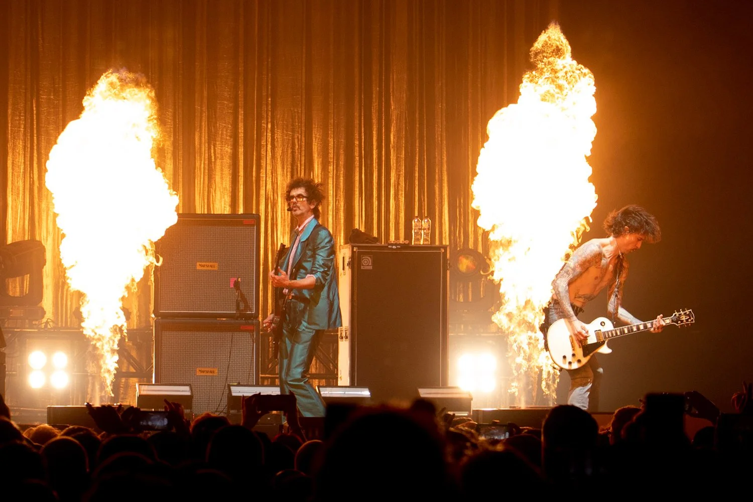 Musicians performing on stage with flames shooting up behind them, gold curtain in background, audience in front.