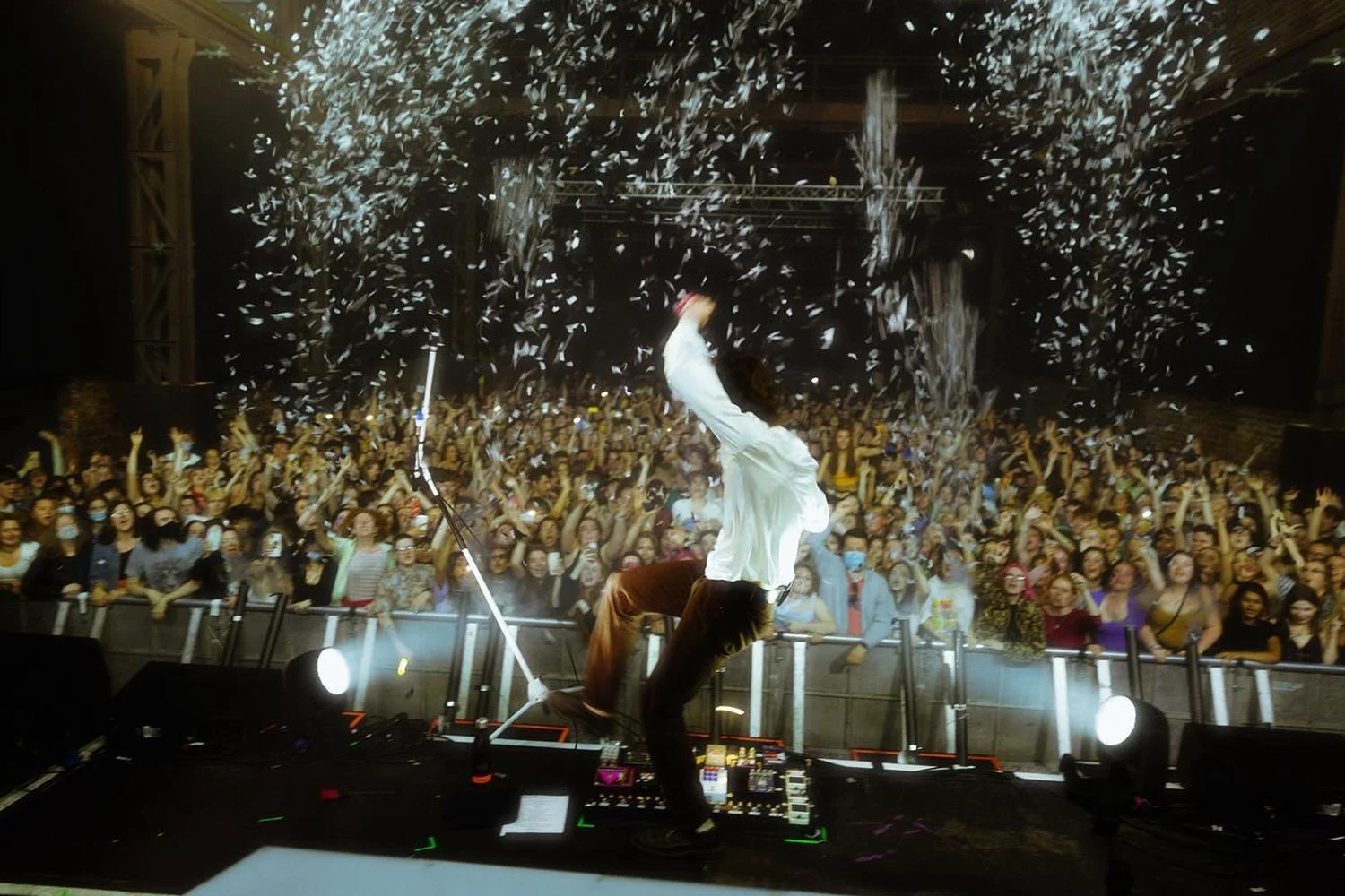 A performer on stage in front of a large crowd, with confetti falling around. The performer is in a dynamic pose, leaning back with one arm raised, and appears to be enjoying the moment. The audience looks excited, many with their hands raised and capturing the moment with their phones.