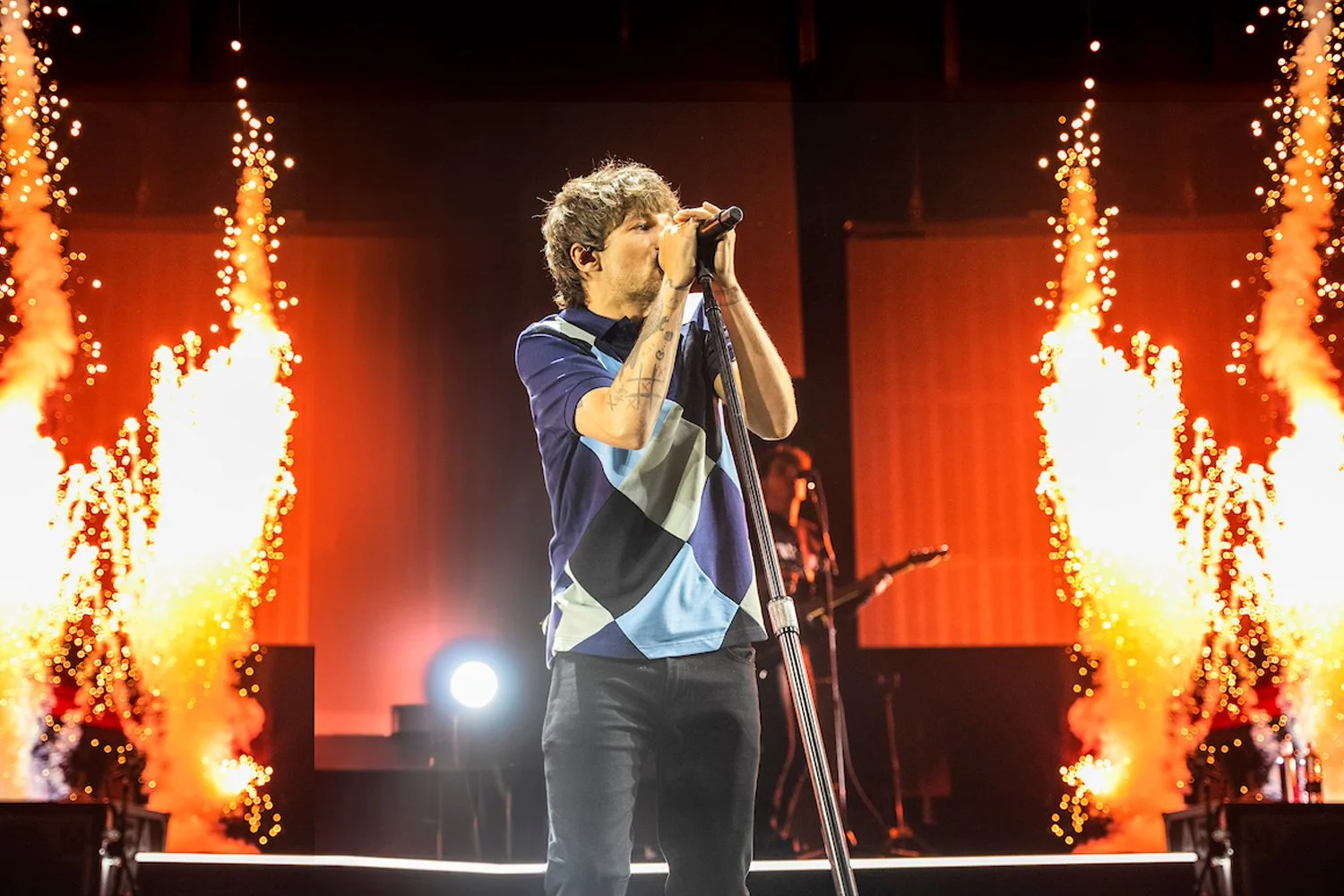 A male singer with tattoos on his arms performs on stage with bright orange flames and pyrotechnics behind him.