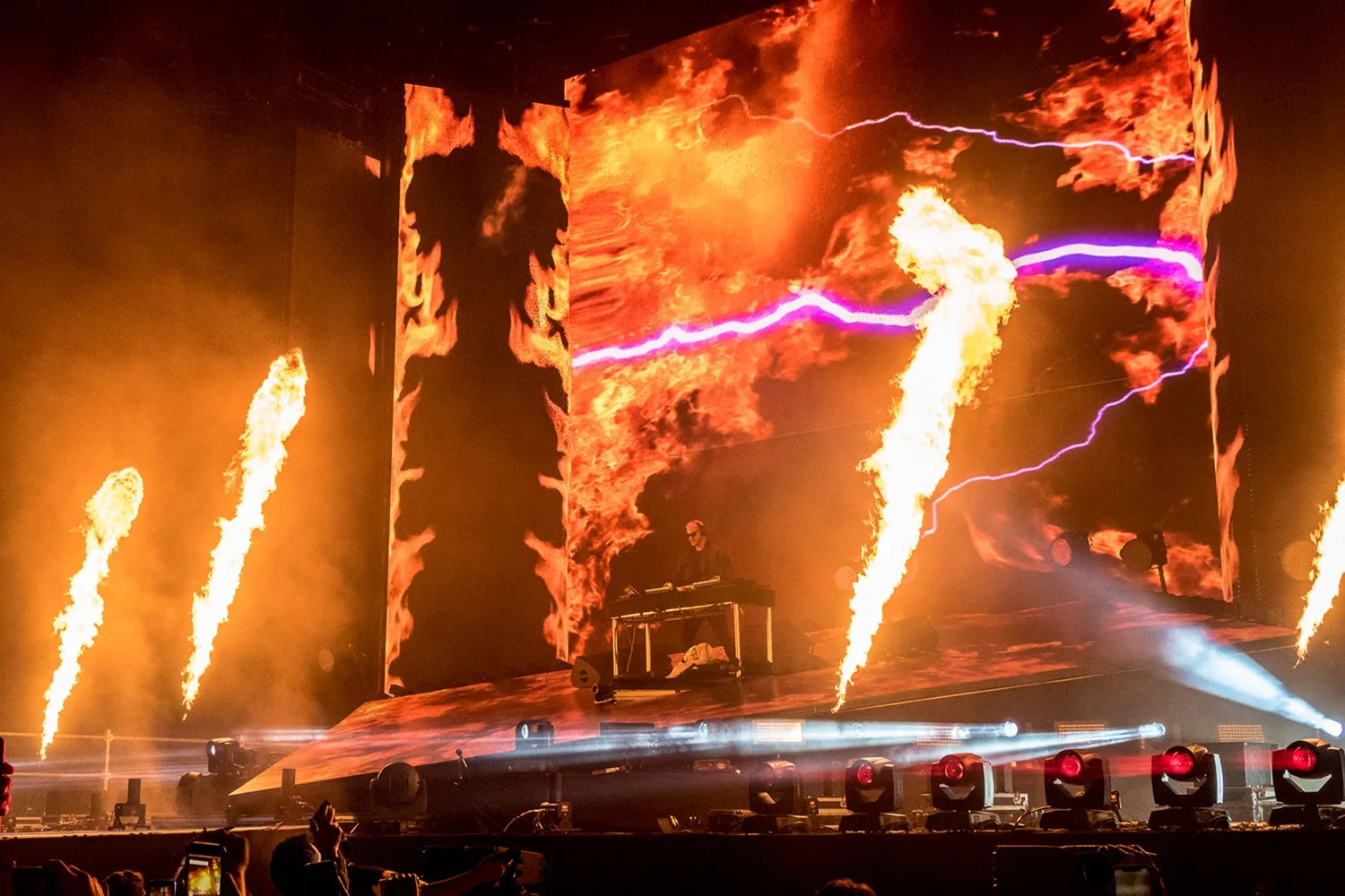 A concert stage with a DJ performing amid flames, fire effects, large digital screens with fiery visuals, smoke, and stage lighting.