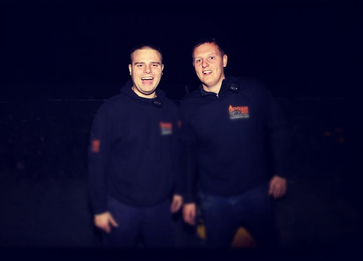 Two young men standing together at night, wearing dark jackets with logos, smiling at the camera.