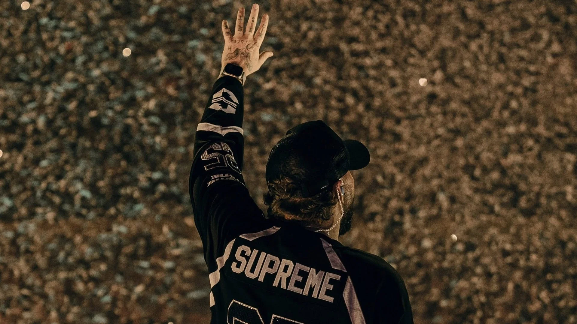 Person with tattoos, wearing a black Supreme jacket and baseball cap, reaching upward with left hand covered in tattoos, in a dimly lit outdoor setting with a gravel ground.
