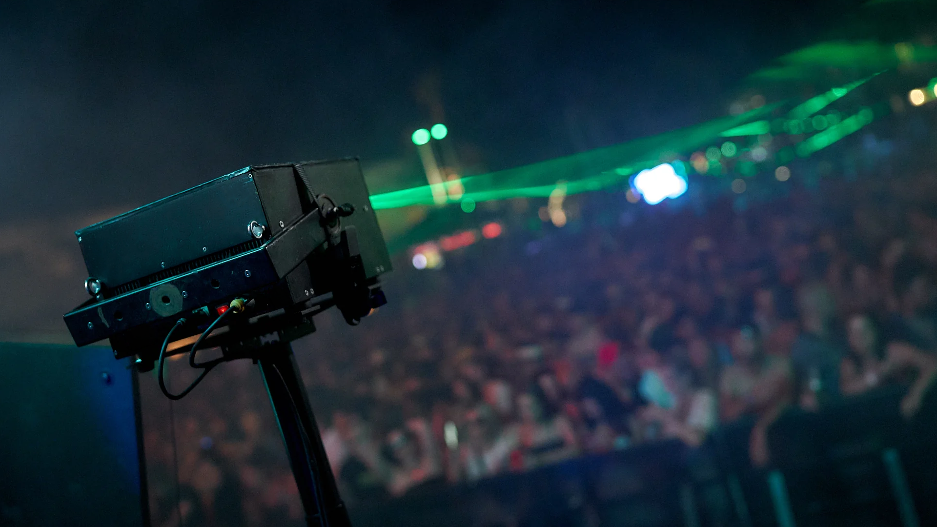 A laser projector emitting green laser beam at night, with a blurred audience in the background.