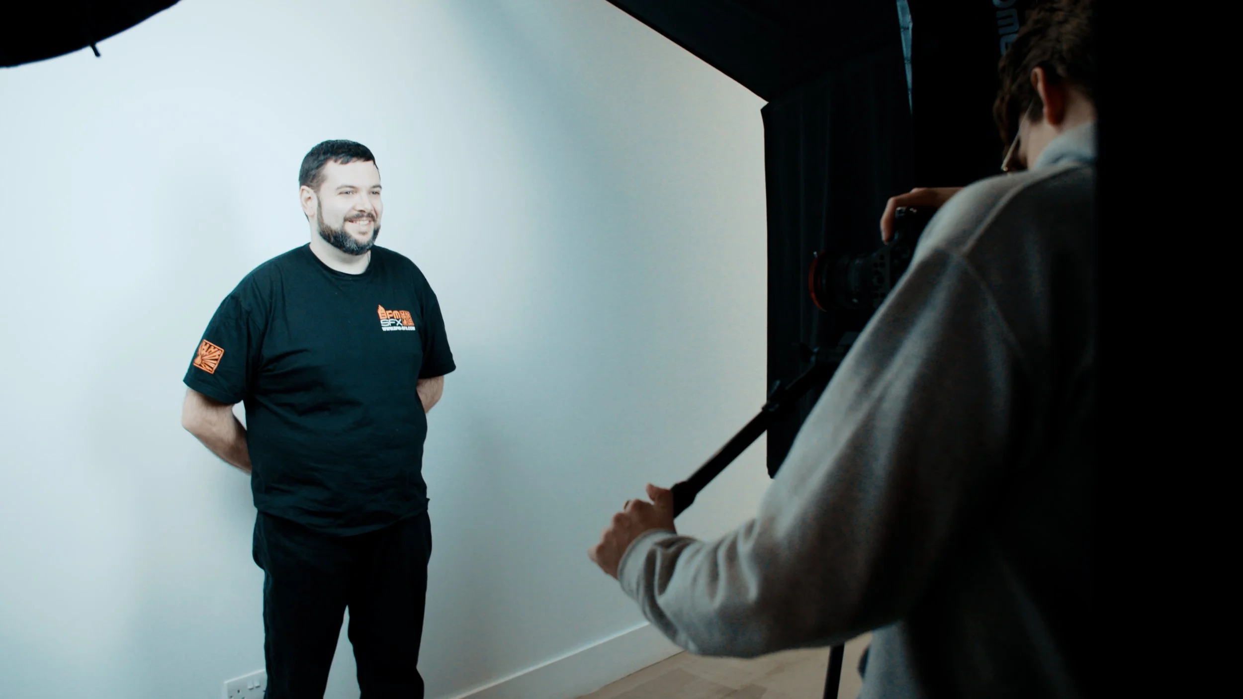 A man with a beard smiling in front of a white screen during a photo or video shoot. Another person is adjusting a camera on a tripod, capturing the shot.