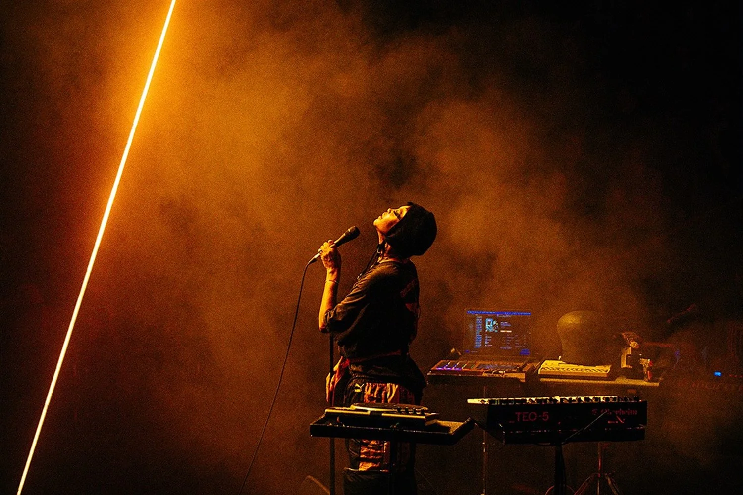 A person with short dark hair, wearing a black jacket, holding a microphone, stands on stage surrounded by electronic music equipment, with orange lighting and smoky atmosphere.